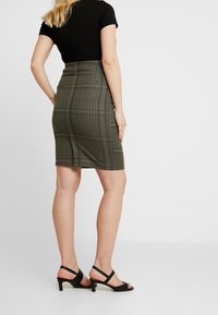 Gonna bodycon verde oliva con pattern a quadri neri. Lunga fino al ginocchio, con un design aderente. Indossata con tacchi neri a strappo.
