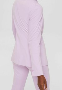 Costume ajusté violet clair comprenant un blazer structuré et un pantalon cintré. Le blazer présente un col cranté et des boutons décoratifs sur les manches.