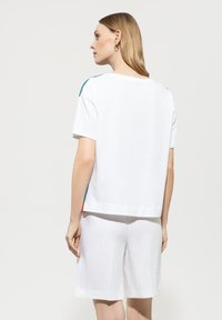 Weißes Baumwoll-T-Shirt mit geradem Schnitt, das blaue und rote Akzente an den Schultern aufweist. Kombiniert mit weißen Leinen-Shorts.