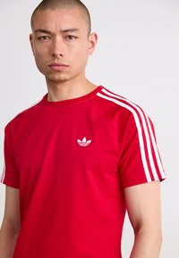 Červené bavlněné tričko s bílými rukávy se třemi pruhy a malým bílým logem na prsou. Krátké rukávy a kulatý výstřih.