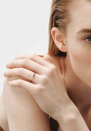 Un anillo de oro con una piedra preciosa verde adorna un dedo esbelto. La mano reposa sobre un hombro desnudo contra un fondo neutro, destacando el anillo.