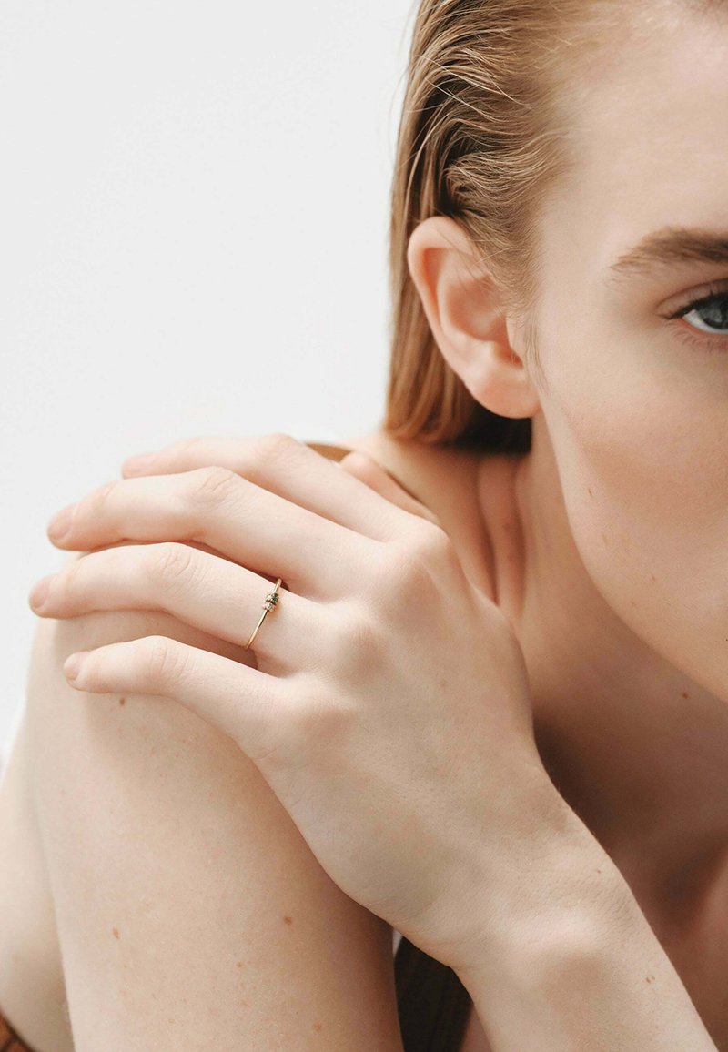 Un anillo de oro con una piedra preciosa verde adorna un dedo esbelto. La mano reposa sobre un hombro desnudo contra un fondo neutro, destacando el anillo.