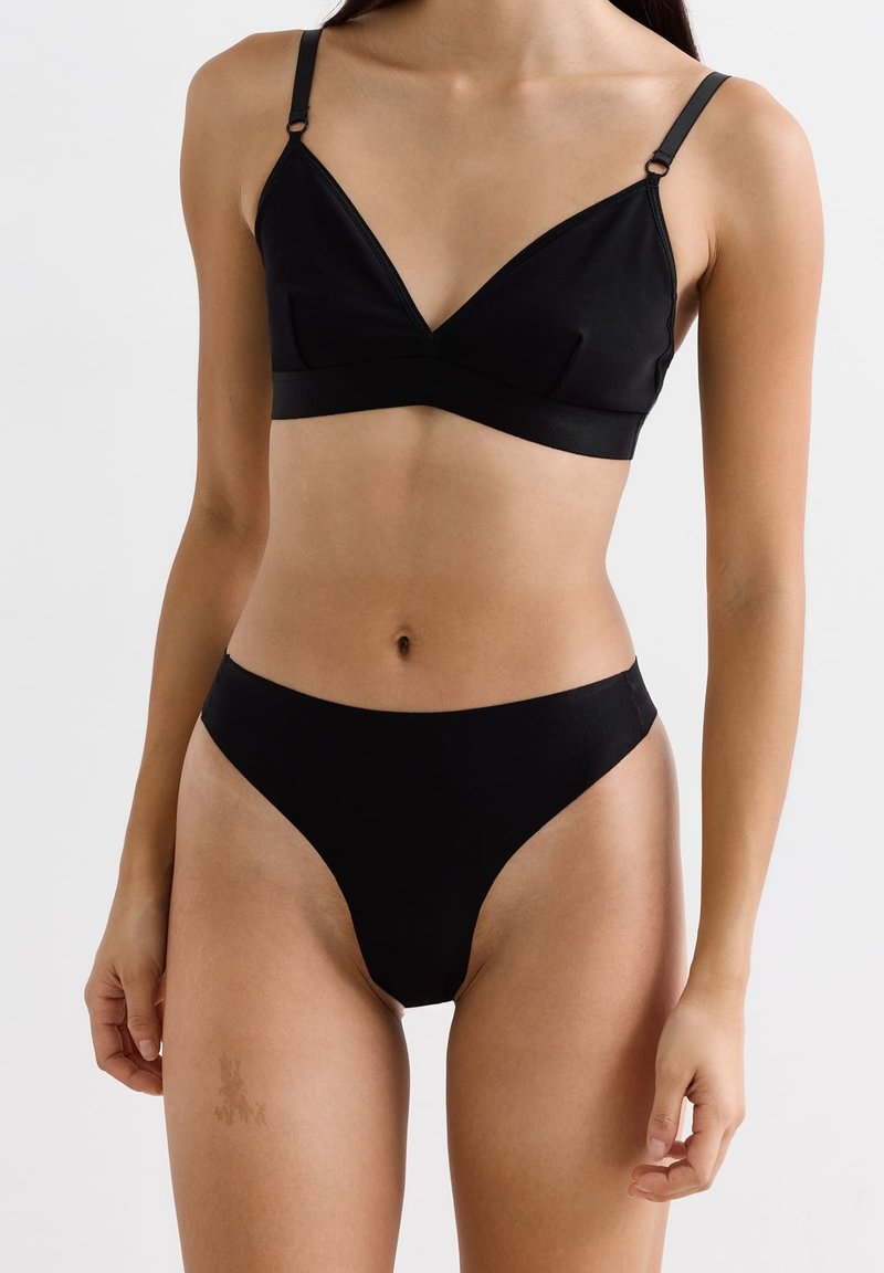 Femme portant un soutien-gorge triangle noir et un bas taille haute assorti, se tenant devant un fond blanc uni.