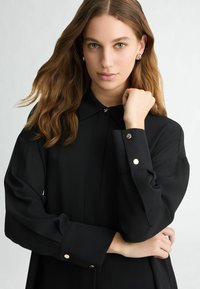 Black blouse with a classic collar, long sleeves, and button accents. Smooth fabric with a relaxed fit and subtle sheen.