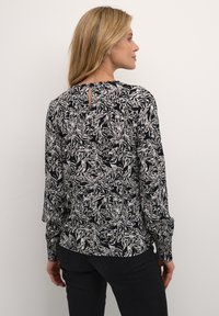 Blusa a maniche lunghe nera con motivo floreale bianco, caratterizzata da un dettaglio a chiave sulla schiena e polsini con bottone. Tessuto morbido e vestibilità comoda.