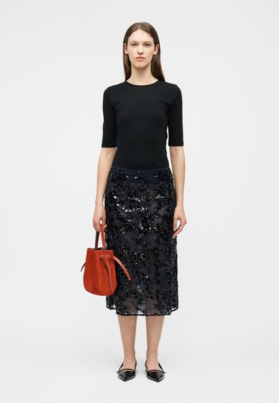 Γυναίκα που φοράει μαύρο εφαρμοστό τοπ, μαύρη μίντι φούστα με παγιέτες, μαύρα μυτερά παπούτσια, κρατώντας μια κόκκινη τσάντα χειρός, μπροστά σε απλό φόντο.