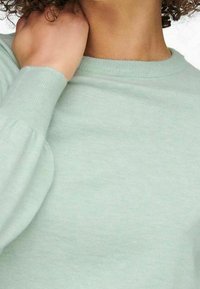 Acercamiento de una persona que lleva un suéter de manga larga verde menta, con una mano tocando suavemente su cuello.