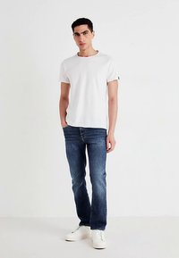 Valkoinen puuvillainen T-paita, jossa on lyhyet hihat, yhdistettynä tummansinisiin farkkuihin ja valkoisiin tennareihin, seisomassa yksinkertaisen taustan edessä.