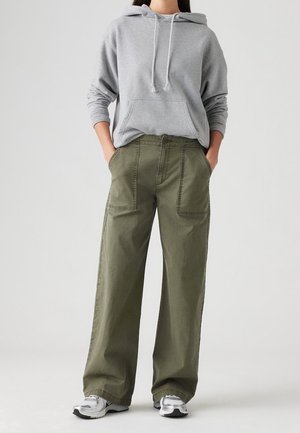 Osoba w szarej bluzie z kapturem, oliwkowozielonych spodniach o szerokich nogawkach z przednimi kieszeniami oraz białych trampkach, stojąca z rękami w kieszeniach.