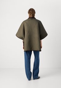 Gequilted olijfgroene poncho met een bruine kraag, brede mouwen en een gestructureerd patroon. Gepaard met losse blauwe jeans en donkere schoenen.