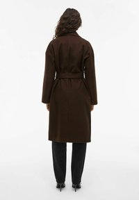 Trench-coat en mélange de laine brun avec une taille ceinturée, un large col et des manches longues, accompagné de pantalons noirs et de chaussures à talons hauts.