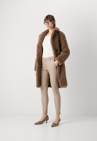 Brun, teksturert faux fur-frakk med krage, satt sammen med tettsittende beige bukser og grå hæler. Modellen bruker briller og en hvit topp.