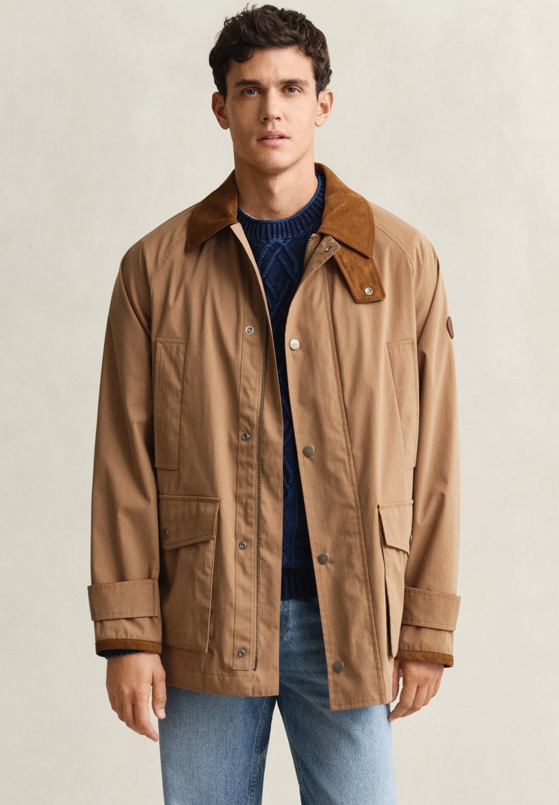 Jeune homme portant une veste marron avec col en velours côtelé et poches avant, par-dessus un pull bleu marine et un jean bleu clair, sur fond uni.