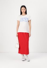 Weißes Baumwoll-T-Shirt mit blauer, blumiger "GAP"-Logoprägung, kombiniert mit einem langen roten Rock. Eine weiße Handtasche und hohe Sneaker runden das Outfit ab.