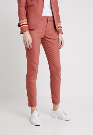 Persoon die strakke broek in roestkleur met gouden en rode manchetdetails draagt, gecombineerd met witte sneakers en een bijpassend colbert.