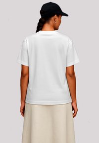 Woman with braided hair wearing a white t-shirt, beige skirt, black cap, and silver hoop earrings, facing away against a plain background.