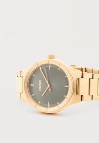 Ρολόι χειρός Hugo σε χρυσό τόνο με μαύρο καντράν, μίνιμαλ δείκτες ώρας και μπρασελέ από μεταλλικούς κρίκους, δείχνοντας ώρα 7:55:02.