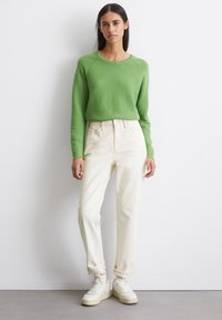 Pull vert à col rond, jeans blancs taille haute et baskets blanches. La tenue présente une coupe décontractée et des poignets retroussés sur le pantalon.