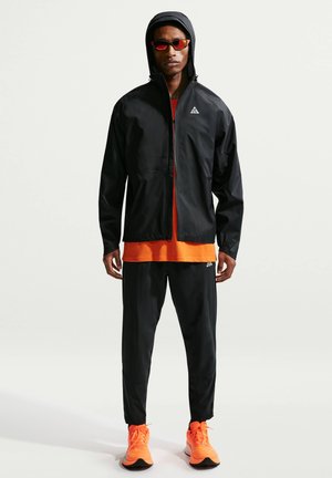 Hombre con chaqueta negra con capucha, pantalones negros, camiseta naranja, zapatillas de deporte naranja brillante y gafas de sol con lentes tintadas de rojo, de pie mirando hacia adelante.