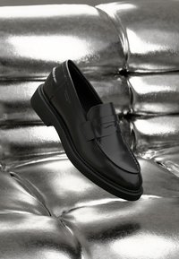Mocassin en cuir noir avec une finition lisse, présentant un détail de sangle à l'avant, un bout rond et une semelle en caoutchouc basse, sur fond métallique.