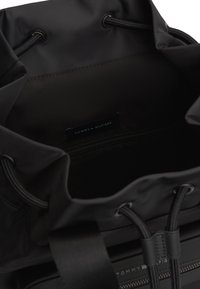 Μαύρη τσάντα νάιλον με κλείσιμο με κορδόνι, διαθέτει εσωτερική τσέπη με φερμουάρ και ετικέτα με λογότυπο. Ομαλή υφή και μινιμαλιστικός σχεδιασμός.