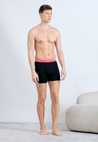 Άνδρας χωρίς πουκάμισο, φορώντας μαύρα εσώρουχα Calvin Klein με κόκκινη μέση, στέκεται ξυπόλυτος σε χαλί κοντά σε μια μπεζ στρογγυλή καρέκλα.