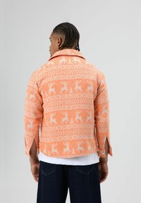 Oranje overhemd met patroon van witte herten en bomen, gemaakt van een gestructureerde stof. Het heeft een kraag, opgerolde mouwen en een afgeronde zoom.