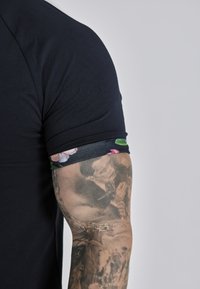 Zwarte T-shirt met korte mouwen met een bloemenpatroon op de manchetten; aansluitende snit; zachte textuur; gecombineerd met gedetailleerde arm tattoos.