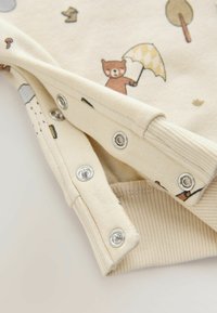 Détail d'un vêtement beige ornée de graphismes animaliers, avec des poignets côtelés et des boutons-pression pour un habillage facile. Tissu en coton texturé.