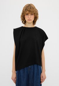 Blusa nera a maniche corte con vestibilità rilassata, caratterizzata da un design asimmetrico e dettagli plissettati sulla spalla, realizzata in un tessuto liscio.