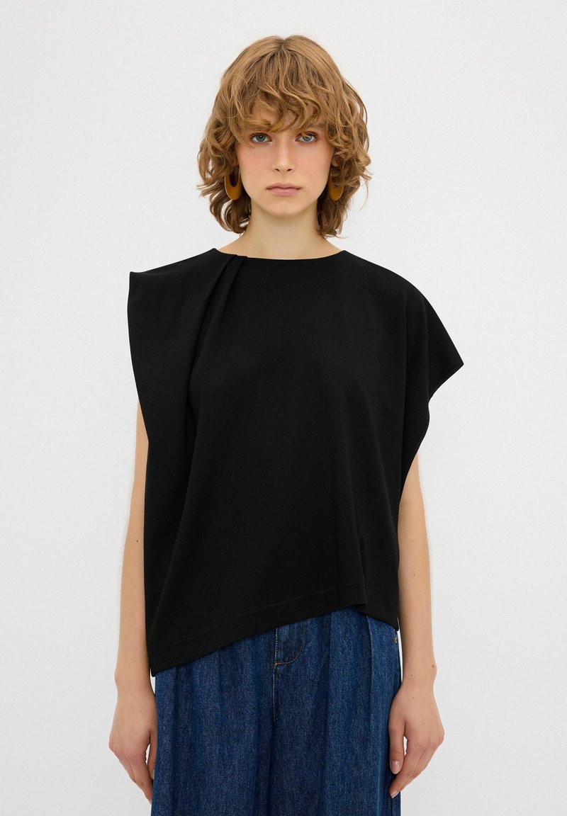 Blusa nera a maniche corte con vestibilità rilassata, caratterizzata da un design asimmetrico e dettagli plissettati sulla spalla, realizzata in un tessuto liscio.