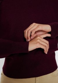 Osoba ubrana w ciemnoczerwony sweter z długim rękawem i beżowe spodnie, z rękami delikatnie skrzyżowanymi na poziomie talii.