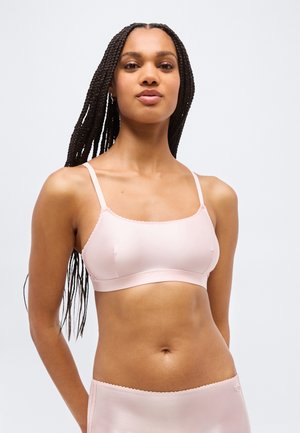 Mujer con largas trenzas, vestida con un bralette rosa pálido y ropa interior a juego, de pie frente a un fondo gris claro.