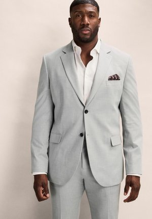 Traje gris claro con dos botones, solapas muesca y bolsillos frontales. Camisa blanca por debajo; el pañuelo de bolsillo tiene un patrón geométrico marrón.