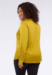 Langärmlige Bluse in Senfgelb, aus schimmerndem seidigem Stoff. Mit klassischem Kragen und Knopfmanschetten. Glatte Textur.