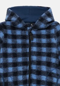 Mėlynos ir juodos languotos zip-up džemperis, pagamintas iš švelnios, tekstūruotos vilnos. Yra su gaubtu, priekiu užtraukiama ir šoninėmis kišenėmis.