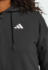 Svart zip hoodie i mjukt material, med vit Adidas-logga på bröstet. Justerbar huva med dragsko och känguruficka.
