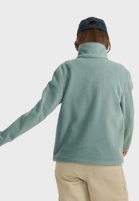 Fleece jacket in light teal, featuring a high collar and long sleeves. Back view shows a smooth texture and relaxed fit.