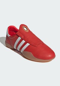 Crvene kožne cipele s tri bijele pruge, okruglim vrhom i detaljem u obliku petlje na klapni. Teksturalni gornji dio i glatki gumeni đon u prirodnoj boji.