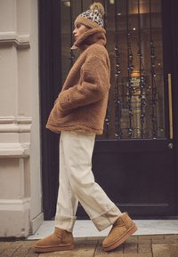 Brun lammeskindsjakke, cremefarvede vidde bukser og beige snørestøvler. Modellen bærer en mønstret strikket hue med pompon på toppen.