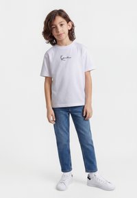 Niño con cabello castaño hasta los hombros, vistiendo una camiseta blanca con diseño gráfico, jeans azules y zapatillas blancas, de pie sobre un fondo blanco liso.