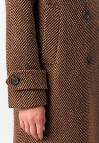 Brun og svart herringbone ullfrakk erme med en knappet mansjett og svarte knapper, som viser en avslappet håndposisjon.