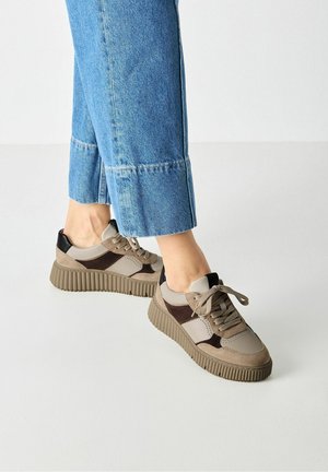 Person iført beige og brune snørestøvler med platform med blå cropped denim jeans på hvidt gulv.