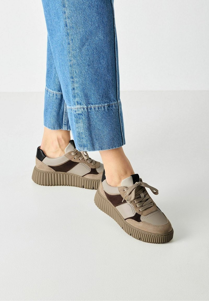 Personne portant des baskets à plateforme beiges et marron à lacets avec un jean en denim bleu raccourci sur un sol blanc.
