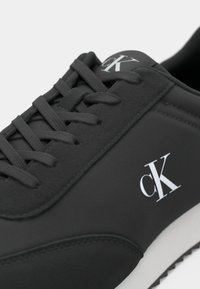 Black athletic shoe with synthetic upper, featuring laces and a white logo. Smooth texture and rounded shape, with a contrast white sole.