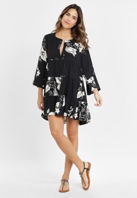 Schwarzes Blumenkleid mit weißen Mustern, gestuftem Design und Glockenärmel. Schlüsselbund-Ausschnitt und weiche Textur. Kombiniert mit braunen Sandalen.