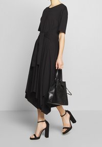 Svart knälång klänning med korta ärmar, asymmetrisk fåll och draperad midjeband. Modell som bär en svart mockaväska och har på sig klacksandaler.