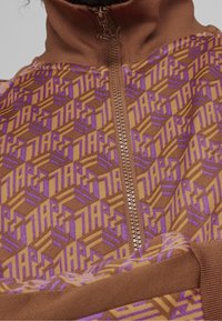 Veste marron à fermeture éclair avec un motif géométrique en violet et jaune. Tissu texturé avec des manches longues et un col haut. Détail prononcé de la fermeture éclair.