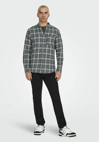 Schwarz-weiß kariertes Flanellhemd mit langen Ärmeln und Knopfleiste, mit einer Brusttasche, getragen mit schwarzen Jeans und Turnschuhen.