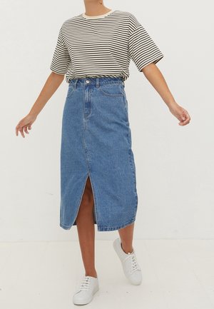 Persoon met een gestreept shirt met korte mouwen, een blauwe denim rok met hoge taille en een voorste split, en witte sneakers, staand op een witte vloer.